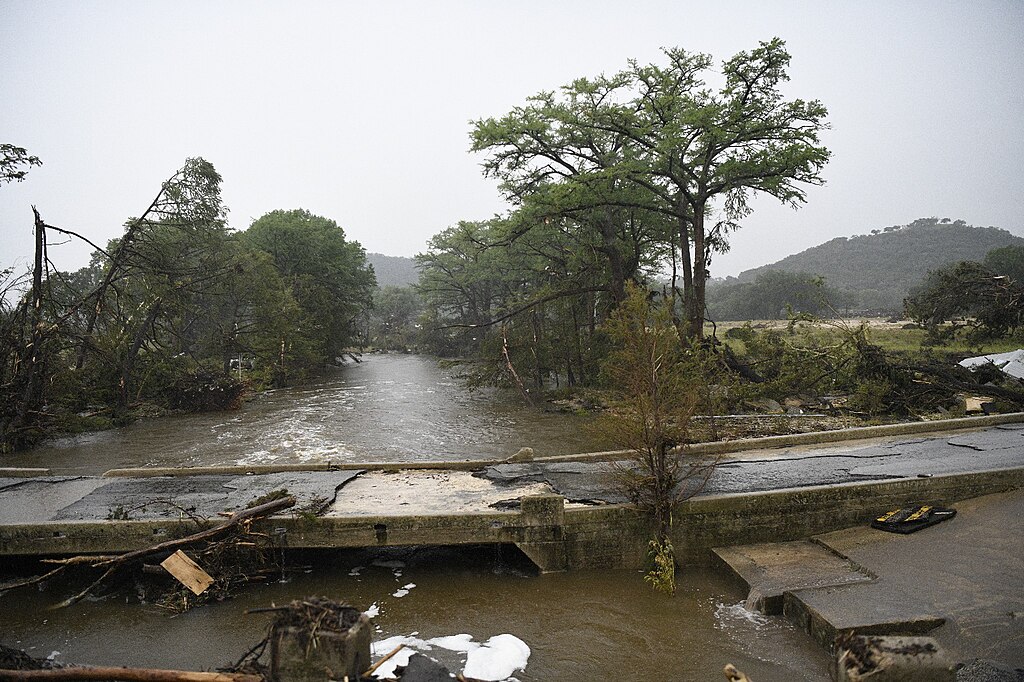 Floodwaters submerge a broken roadway surrounded by downed trees and debris in a rural Texas community after severe flooding.