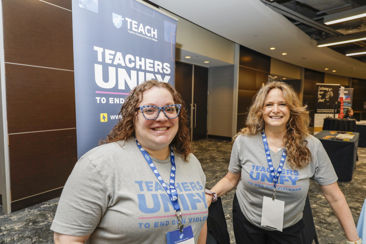 Sarah Lerner and Abbey Clements smile in front of a 'Teachers Unify to End Gun Violence' banner at the AFT TEACH 2025 conference. Both are wearing matching gray Teachers Unify t-shirts and conference lanyards.